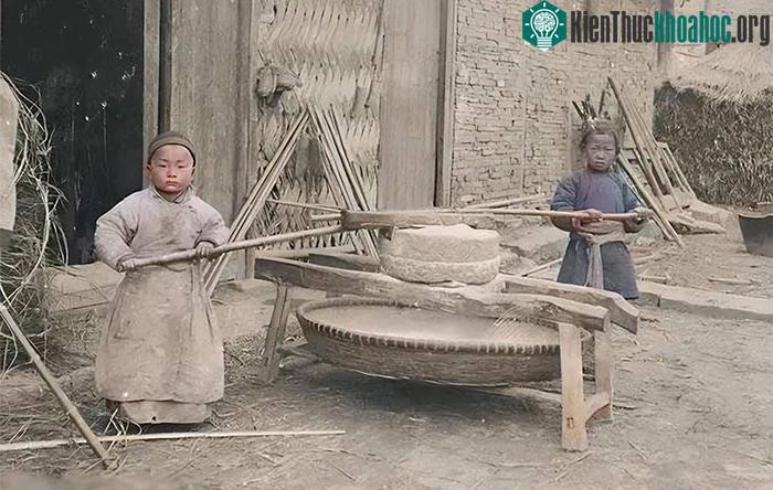 Hai chú bé khoảng sáu, bảy tuổi đang kéo cối xay trước sân nhà