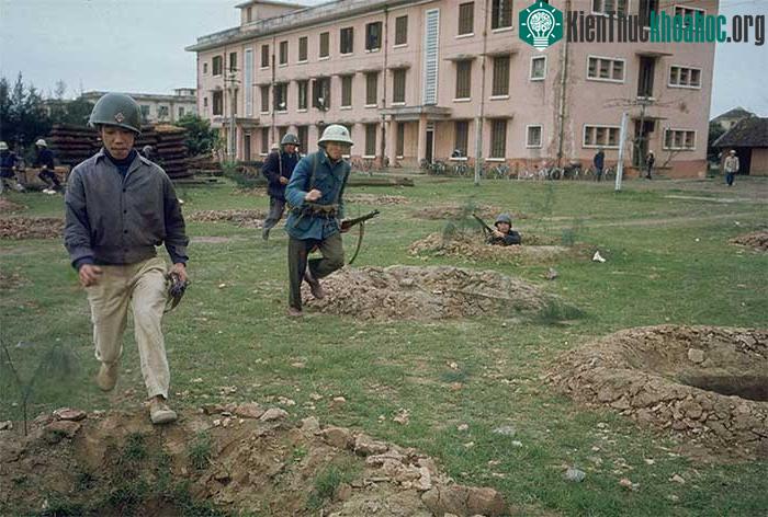 Dân quân Hà Nội luyện tập trên thao trường, phía trước một nhà máy sản xuất xe đạp.